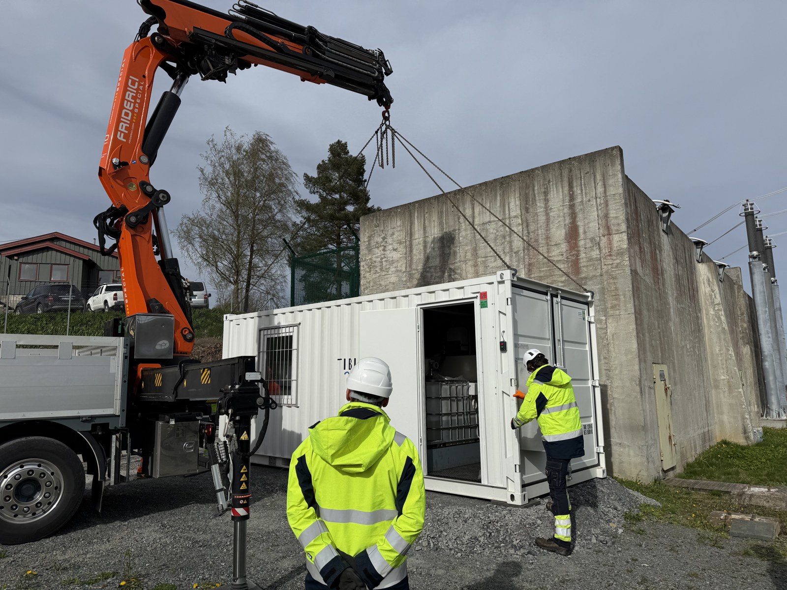 Field technicians opening the doors of a white bionrec treatment container next to a substation wall in Oslo, revealing stacked intermediate bulk containers of bacterial solution inside