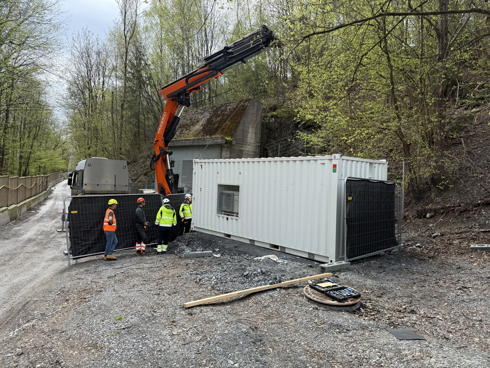 A crane lorry lowering a white treatment container onto a gravel pad beside a wooded access road in Oslo, with four bionrec field technicians in high-visibility clothing standing behind site fencing